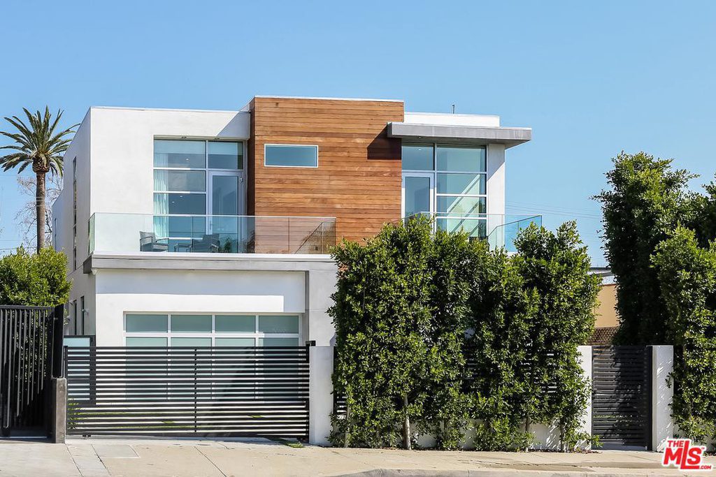 Modern two-story house with large windows, white and wood facade, glass balcony, and tall hedge. The house features a gated driveway and is situated in a sunny location.