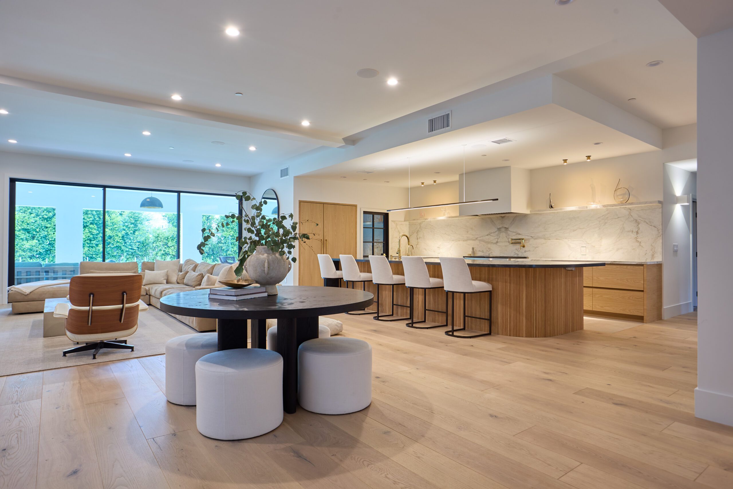 Modern open-concept living area with a kitchen and dining space, featuring light wood flooring, white furniture, a round table with stools, a large kitchen island, and floor-to-ceiling windows.