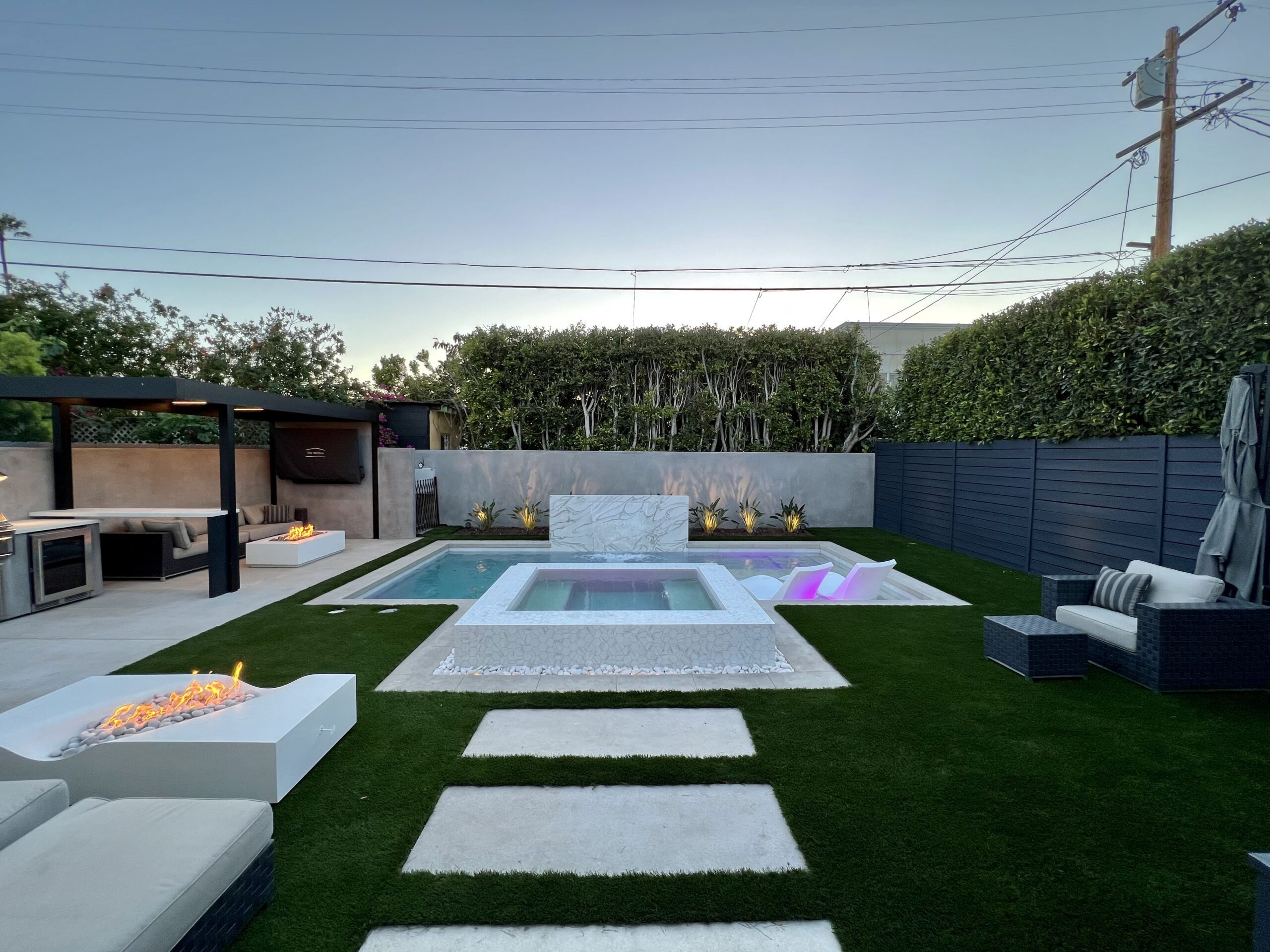 A backyard with a pool and hot tub, surrounded by artificial turf. There's an outdoor seating area with a fire pit and a covered kitchen in the background.