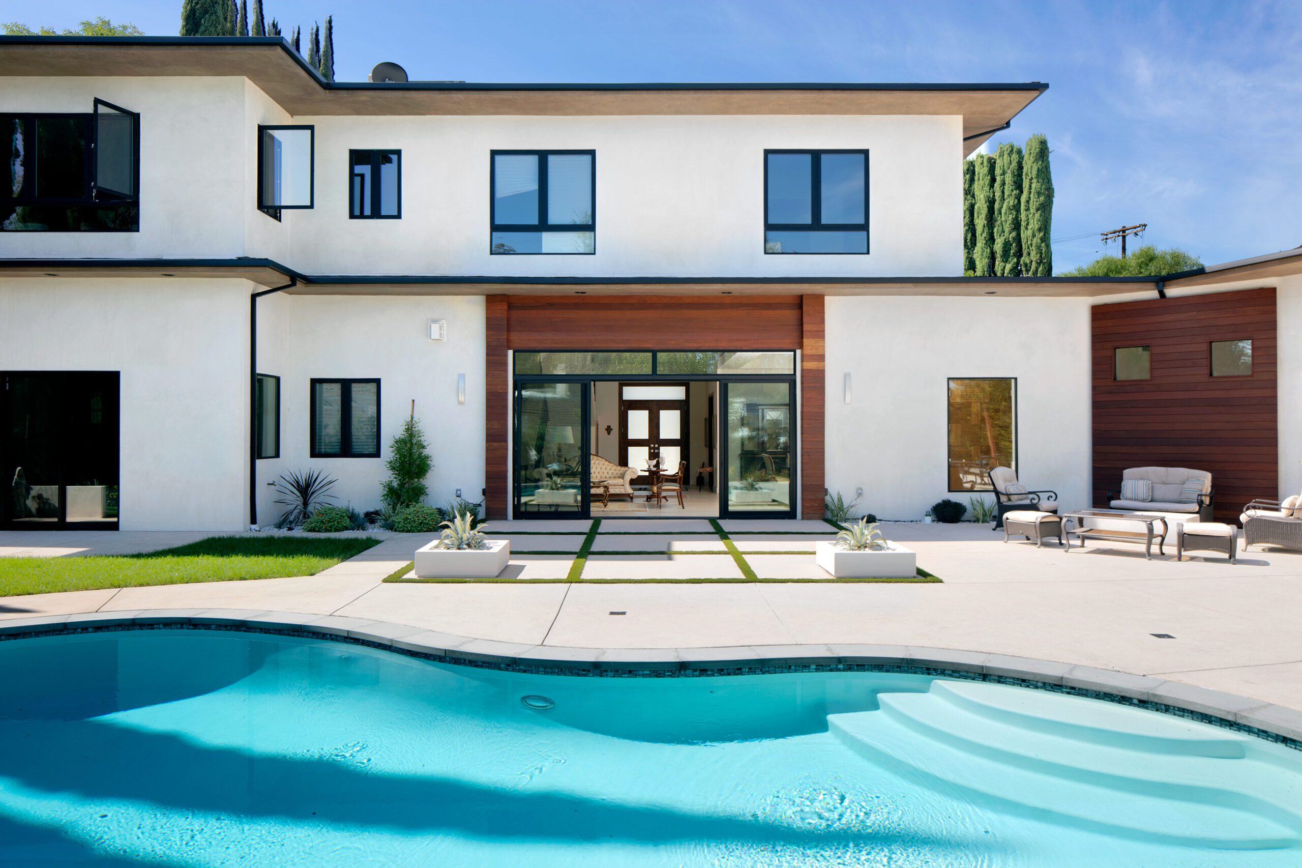 Modern two-story house with large windows, a pool in the foreground, and a spacious patio area with outdoor furniture.