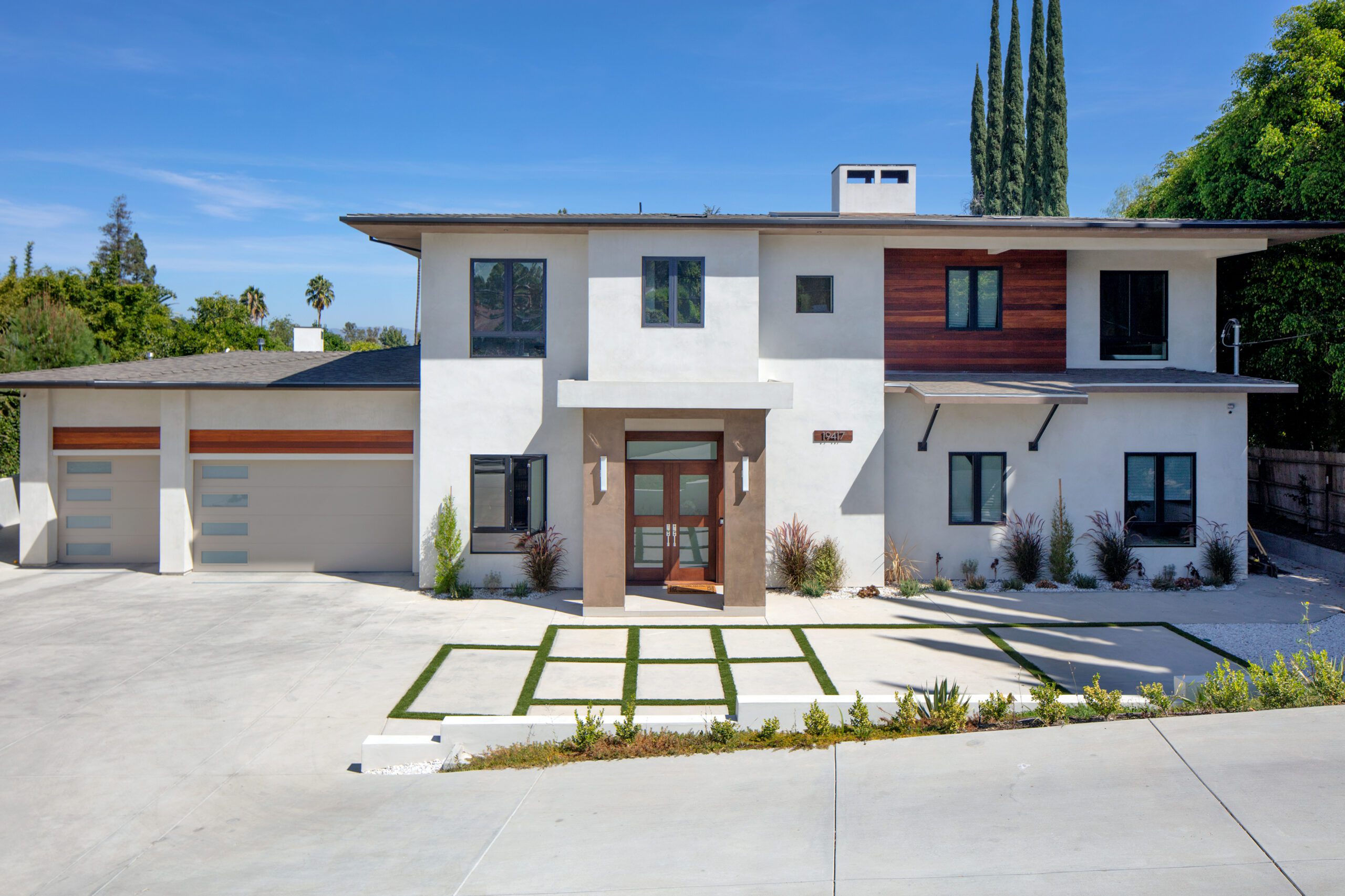 A modern two-story house with a flat roof, large garage, and minimalist landscaping, featuring a mix of white stucco and wooden accents on the exterior.